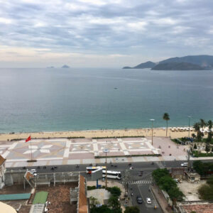 Aerial view of a sandy beach and calm sea with distant islands, palm trees, and a patterned promenade along the shore; city buildings in foreground.