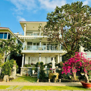 White modern house with glass balconies, lush plants, and a large tree on the right under a blue sky.