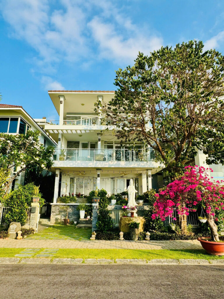 White modern house with glass balconies, lush plants, and a large tree on the right under a blue sky.