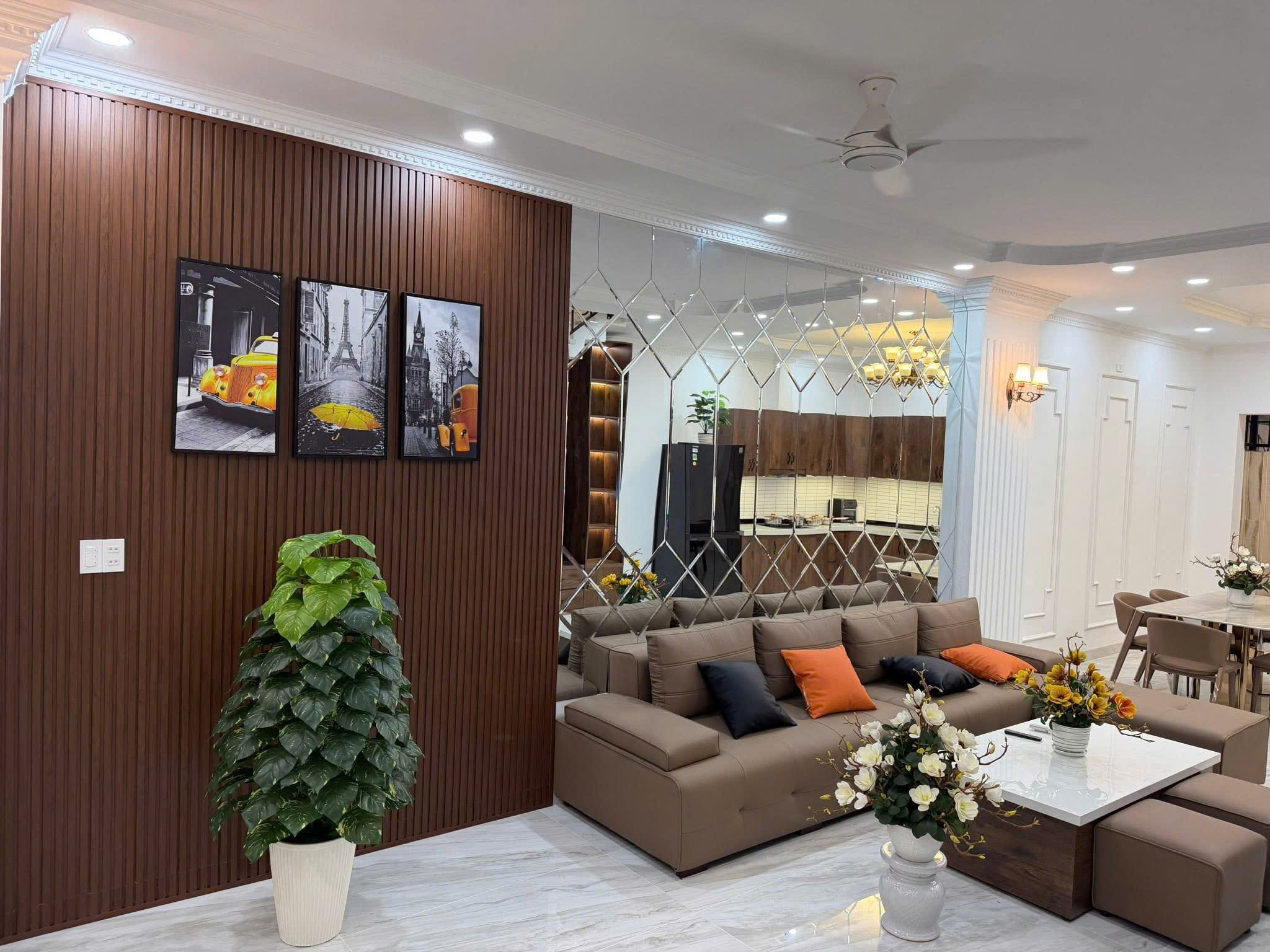 Modern living room with a brown wood slat wall, framed black-and-white art, and a brown sectional with orange cushions.