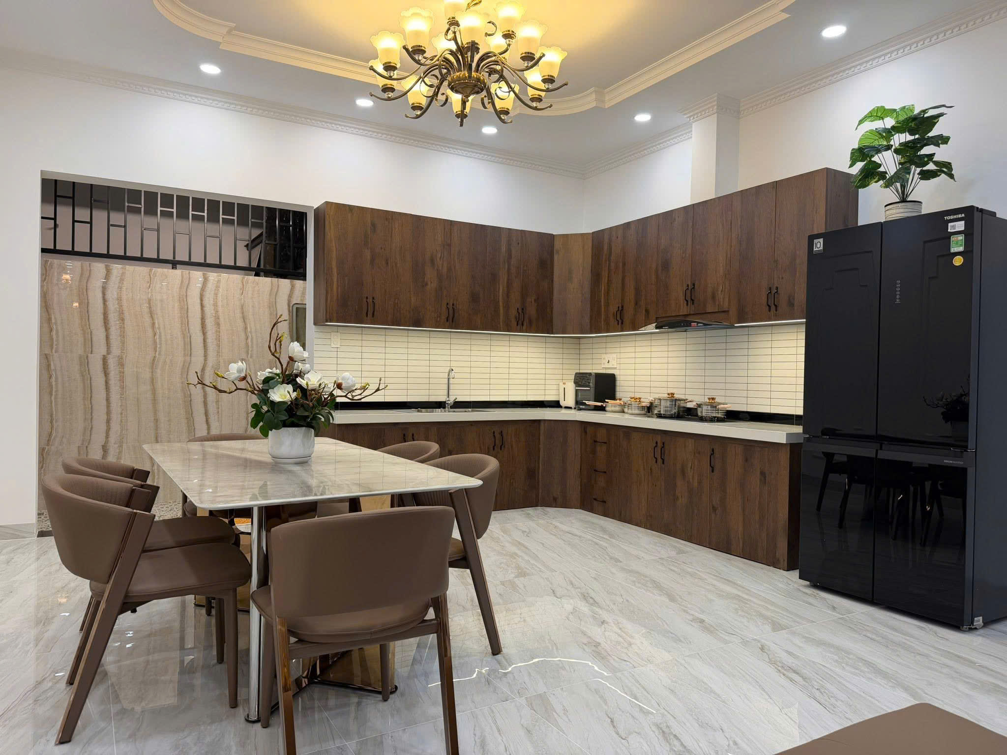 Modern kitchen with brown wooden cabinets, marble dining table, and taupe dining chairs beneath a chandelier; black refrigerator on the right and a potted plant on top.