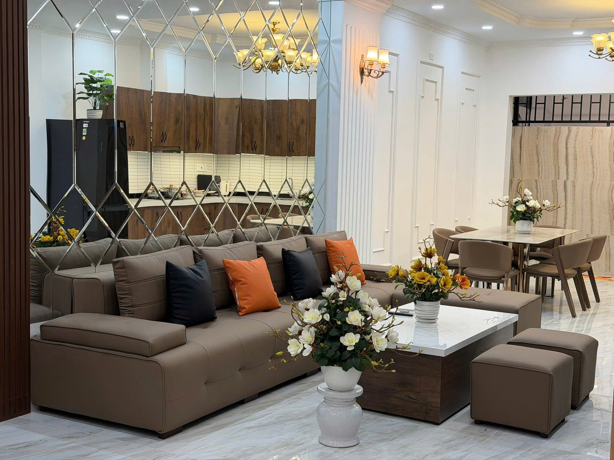 L-shaped brown sectional sofa with orange and black cushions in a bright showroom, mirrored lattice wall, and marble floor vase of white flowers in foreground