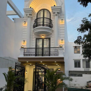 White elegant house facade with ornate arched balcony, black wrought-iron railing, and warm exterior lights at dusk/evening setting.
