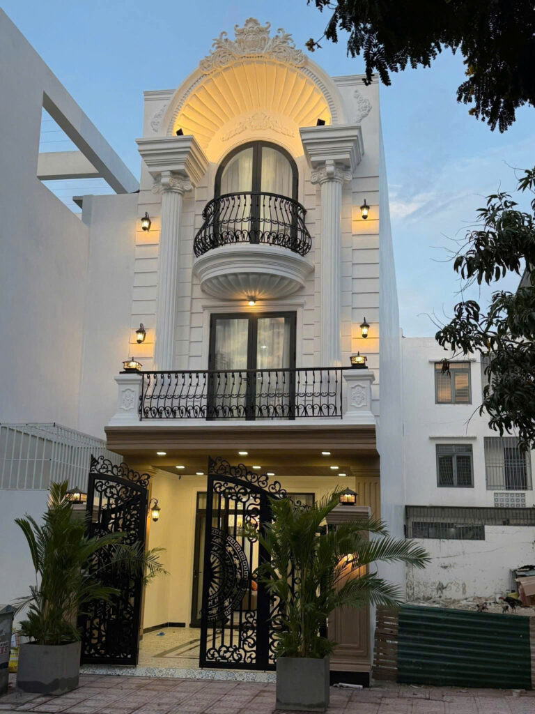 White elegant house facade with ornate arched balcony, black wrought-iron railing, and warm exterior lights at dusk/evening setting.