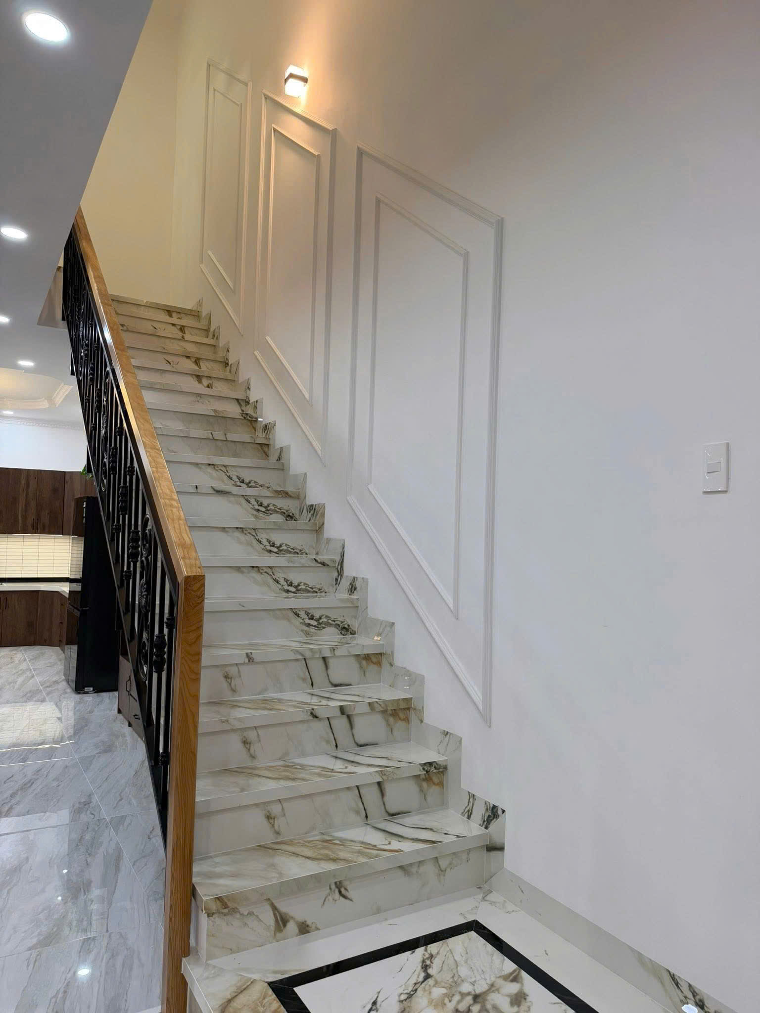Marble staircase with black wrought-iron railing and wooden handrail, ascending beside a white paneled wall in a modern interior.