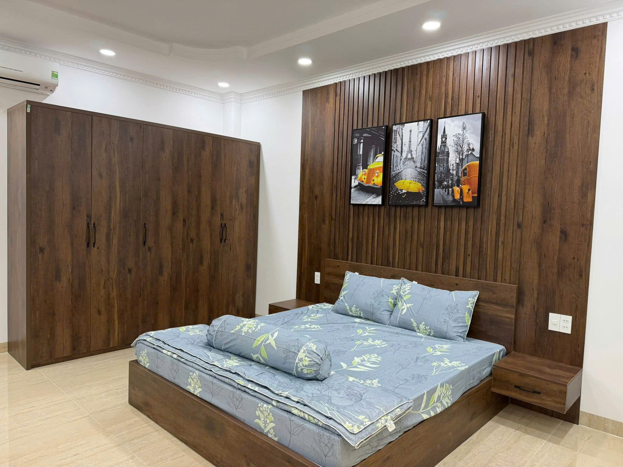 Modern bedroom with dark wood built-in wardrobes, a low platform bed with blue floral bedding, and a trio of black-and-white cityscape prints featuring yellow taxis above the headboard.