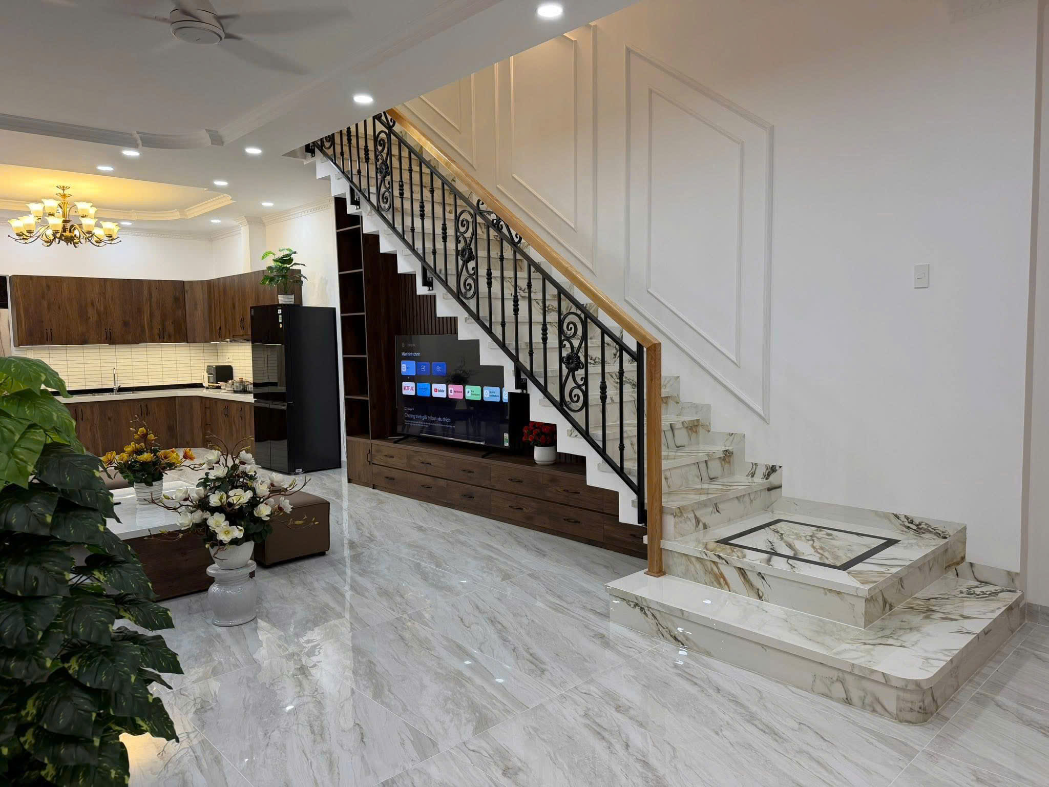 Open-plan home interior with a marble floor, wooden kitchen cabinets, and a decorative staircase with a black wrought-iron railing. A chandelier hangs from the ceiling.