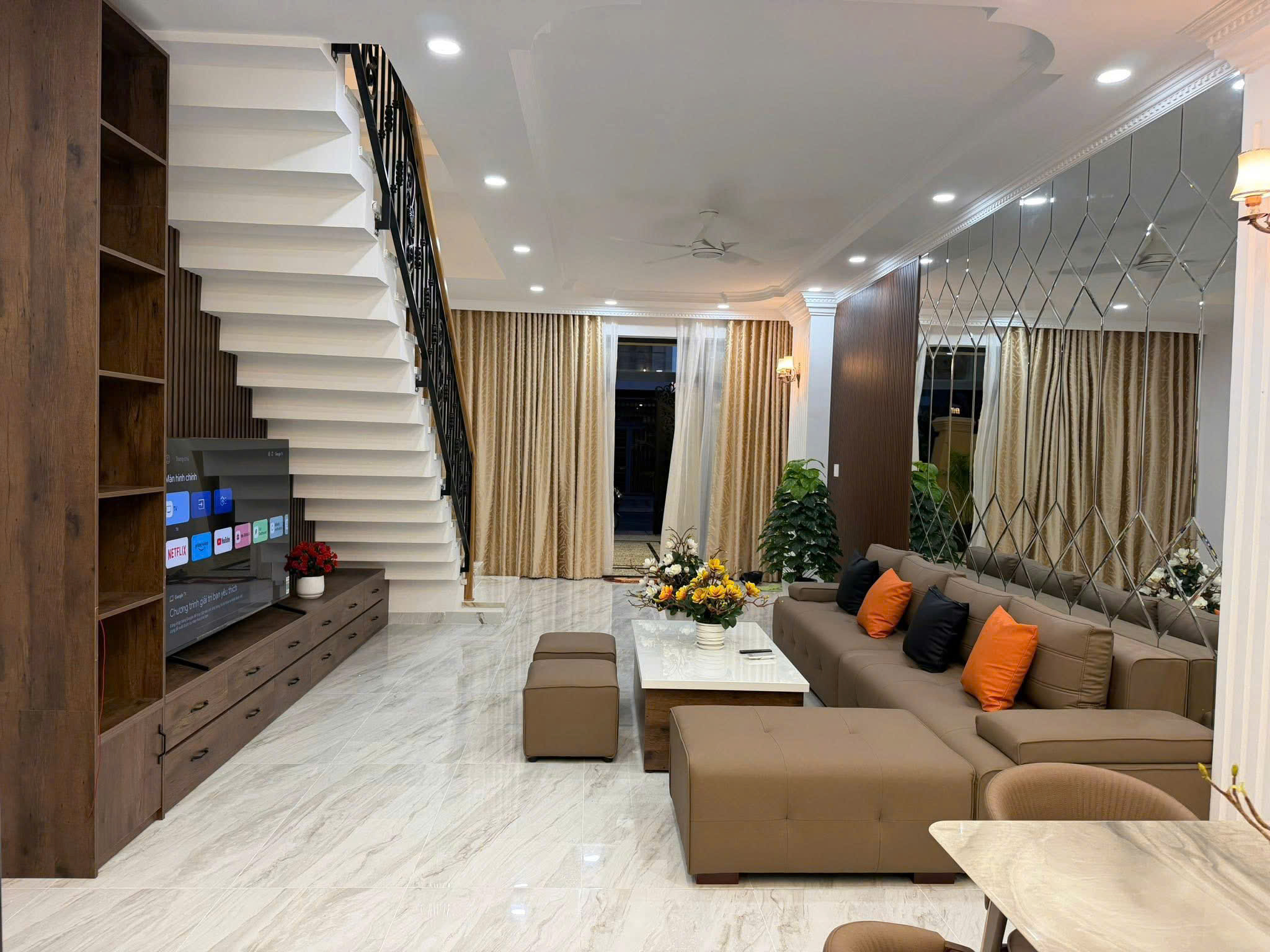Modern living room with white cantilever staircase on left, wood cabinets, TV, beige sectional sofa with orange pillows, marble floor, and a mirrored wall on the right.
