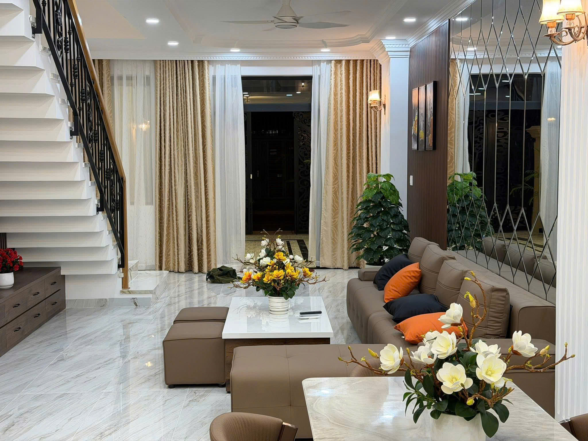Luxurious living room with white marble floors, beige sectional sofa with orange pillows, and glass-topped coffee tables; staircase on the left.