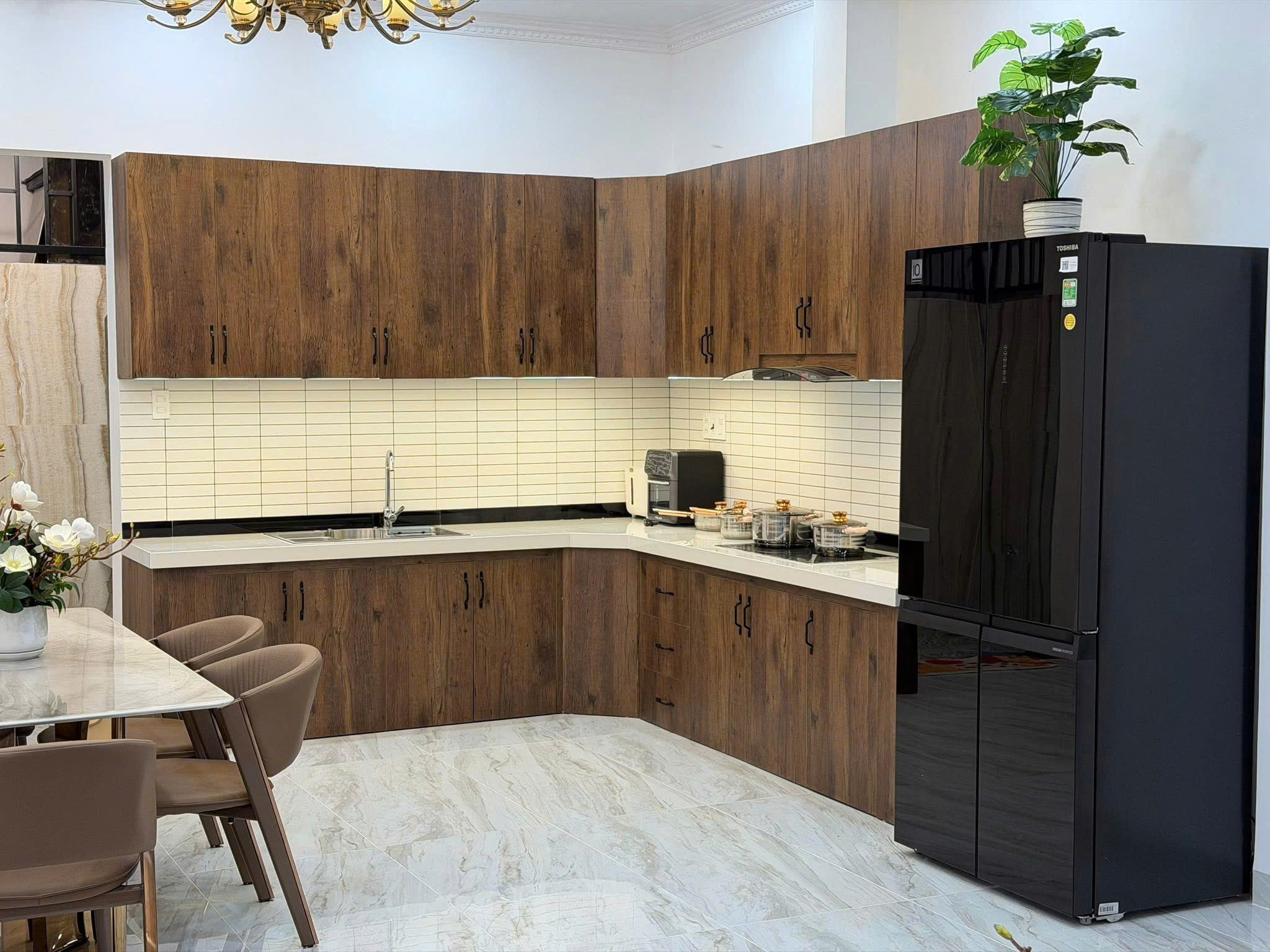 Modern kitchen showroom with dark wood cabinets, white tile backsplash, marble floor, and a tall black refrigerator with a plant on top.
