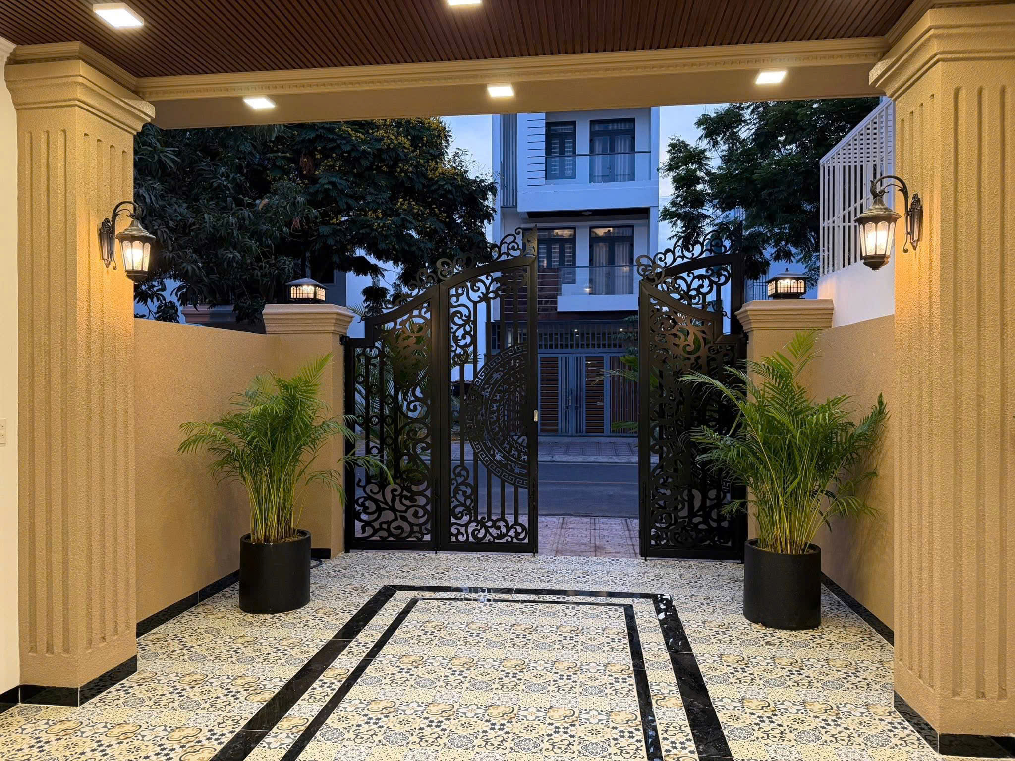 Entrance to a gated courtyard with decorative black wrought-iron gates, flanked by beige columns and potted plants, lamps glowing. (informative)