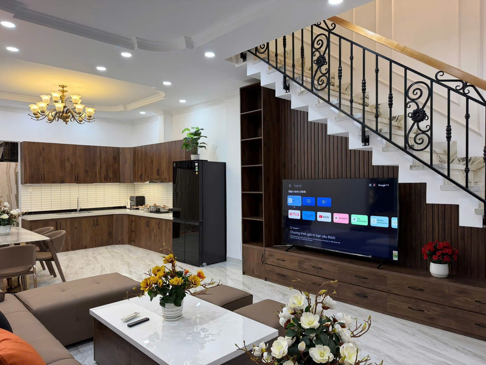 Open-concept living area with dark wood cabinetry, marble countertops, a chandelier, and a staircase with decorative iron railing beside a large TV.