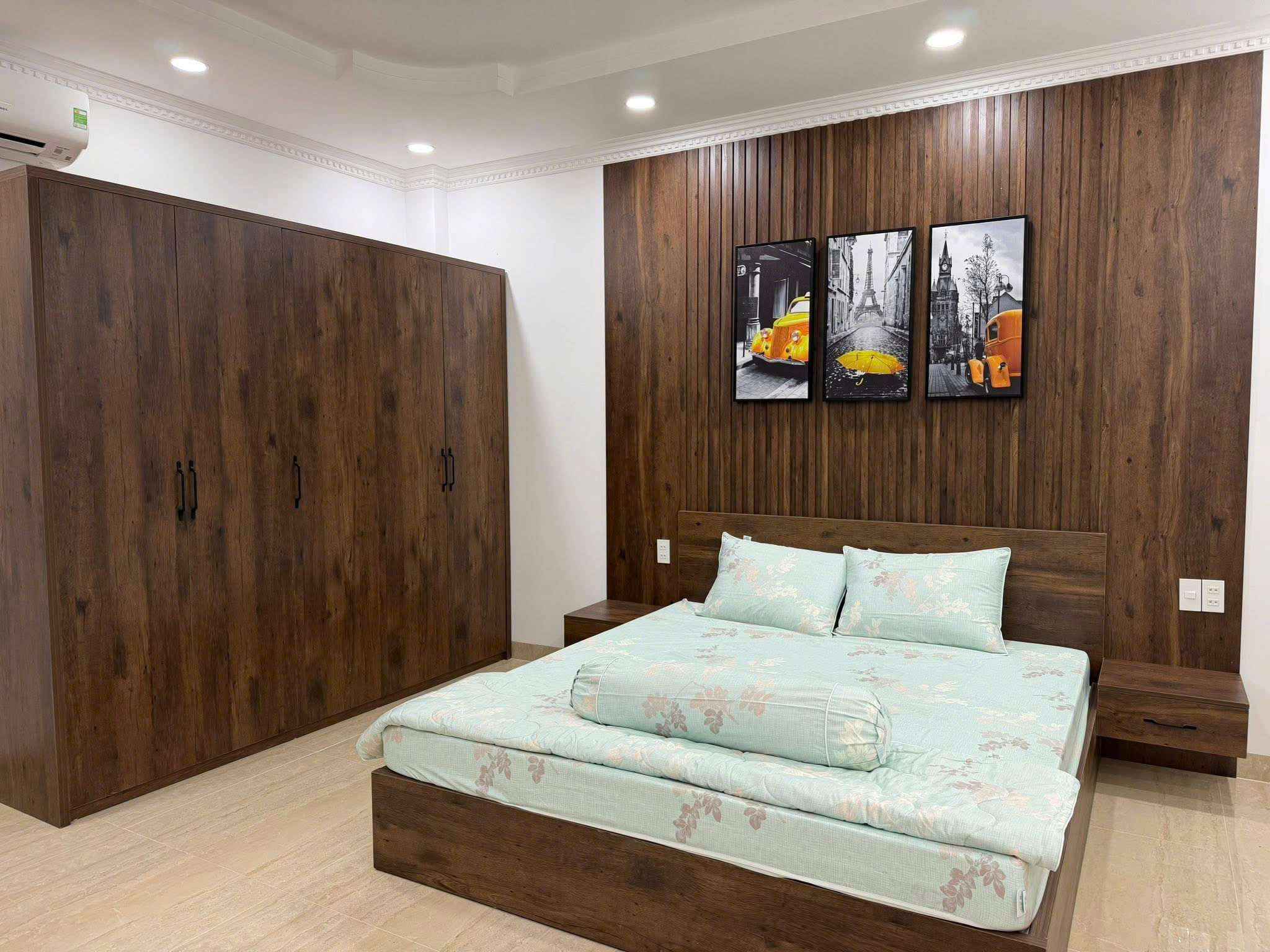Bedroom with dark wood paneled wall and built-in wardrobes; a bed with light blue floral bedding sits beneath a trio of framed black-and-white art prints featuring a yellow taxi, the Eiffel Tower, and a yellow umbrella.