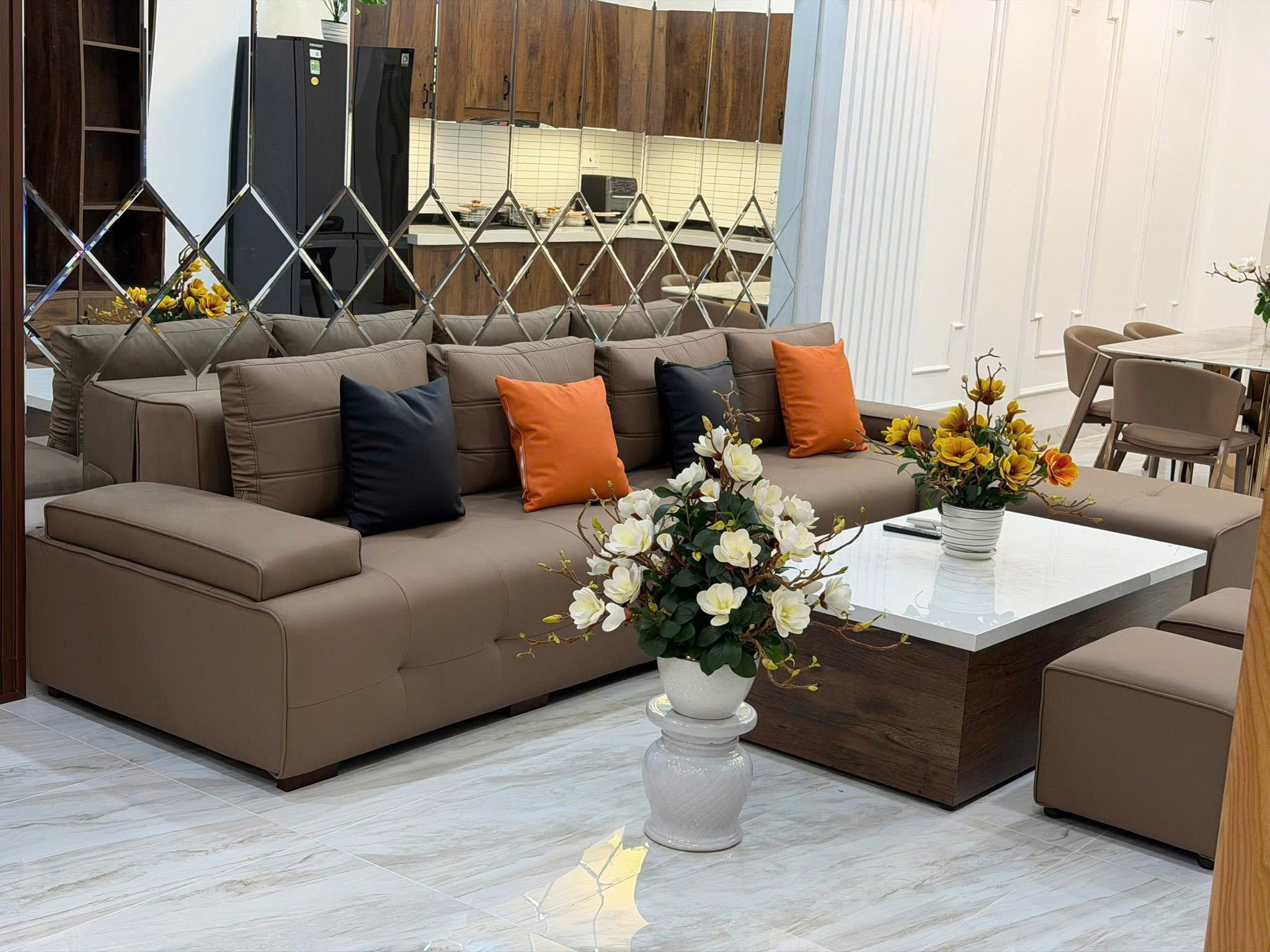 Modern living room with a tan sectional sofa, orange and dark cushions, and a white marble coffee table holding a vase of white and yellow flowers; a mirrored diamond-pattern divider and a kitchen with dark wood cabinets are in the background.
