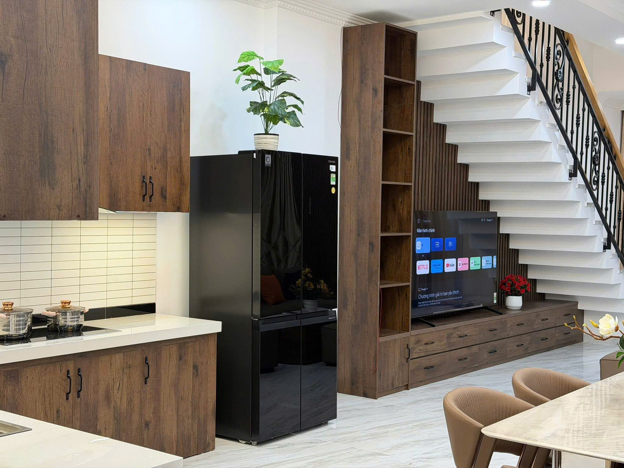 Open-plan kitchen and living area with dark wood cabinets, a black side-by-side refrigerator, and a white staircase with a black railing nearby.