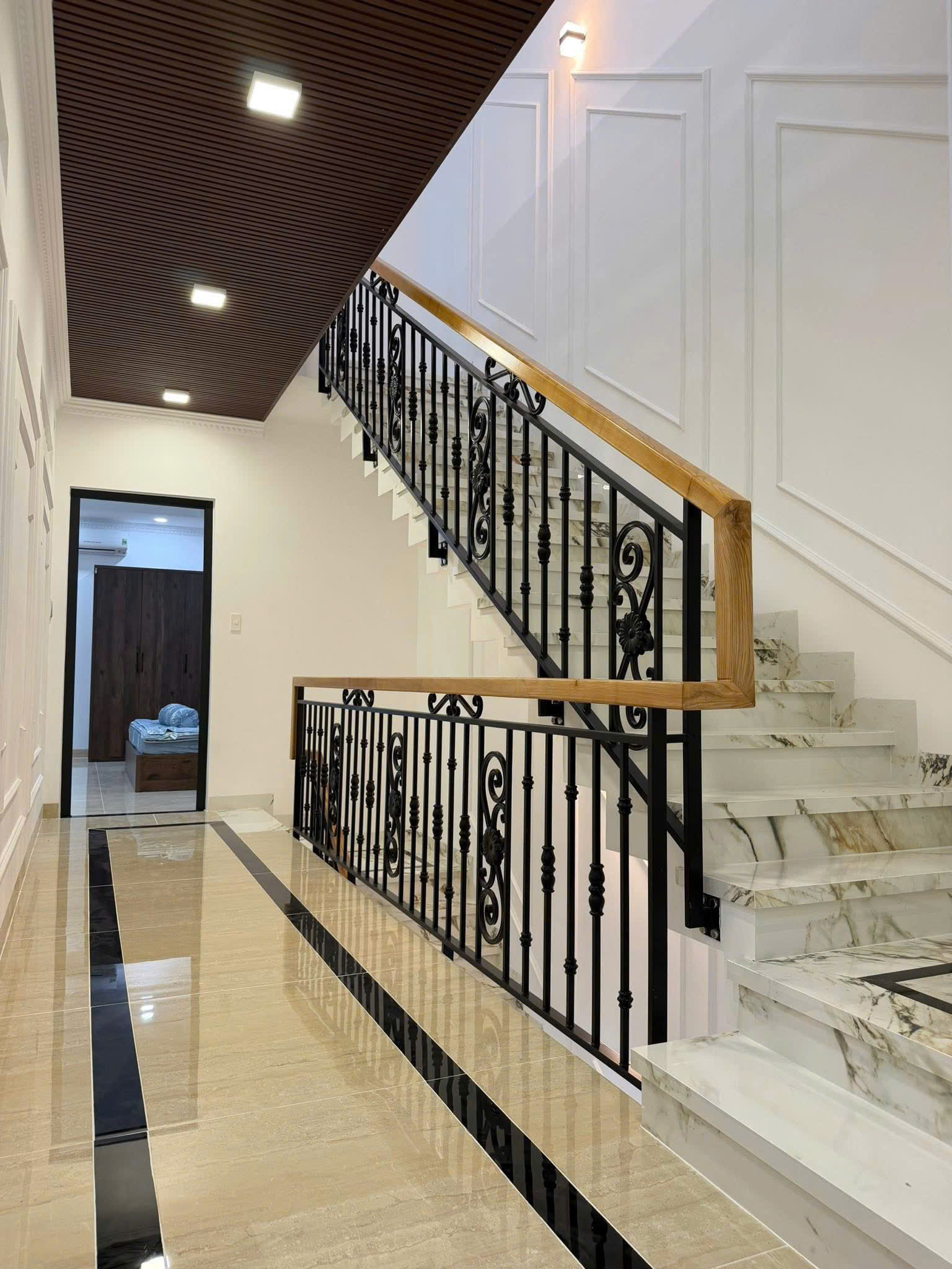 Marble staircase with black wrought-iron railing and wooden handrail in a bright, paneled white interior hallway.