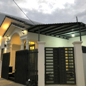 White house with arched entryway, decorative columns, and long black vertical-slatted fence; illuminated porch lights under a metal carport.