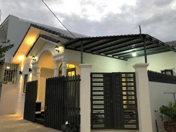 White house with arched entryway, decorative columns, and long black vertical-slatted fence; illuminated porch lights under a metal carport.