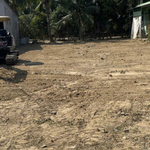 Open dirt lot with tire tracks and palm trees in the background; a small excavator sits on the left.