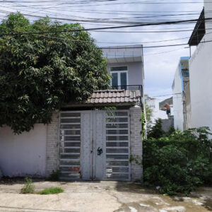 Metal gate with horizontal slats in front of a gray brick wall and a two-story house, partly hidden by a large tree on the left and overhead power lines.