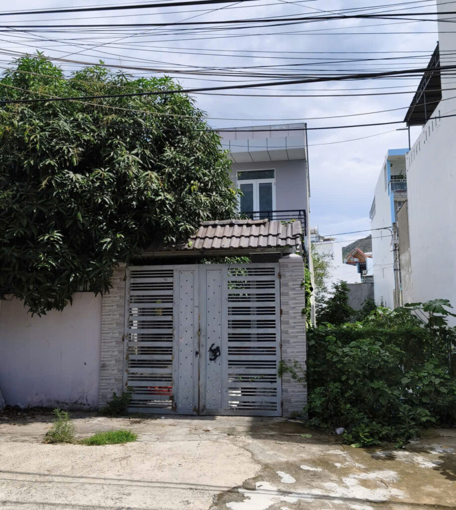 Metal gate with horizontal slats in front of a gray brick wall and a two-story house, partly hidden by a large tree on the left and overhead power lines.