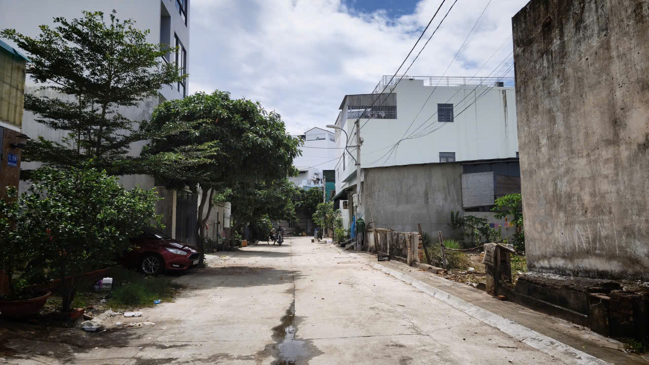 Narrow sunlit urban alley with a parked car on the left, trees, concrete walls, and overhead power lines.
