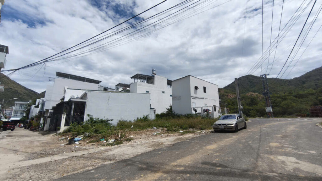 Street view of a quiet residential area with white modern buildings, a parked silver car, and hills in the background under a cloudy sky.