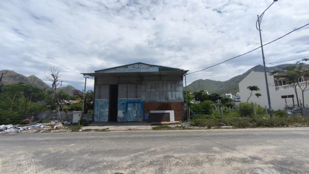 A small, dilapidated warehouse by a road with piles of trash on the left and hills in the distance, under a cloudy sky.