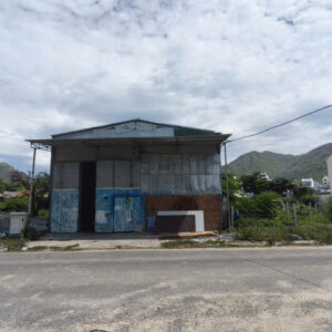A small, dilapidated warehouse by a road with piles of trash on the left and hills in the distance, under a cloudy sky.