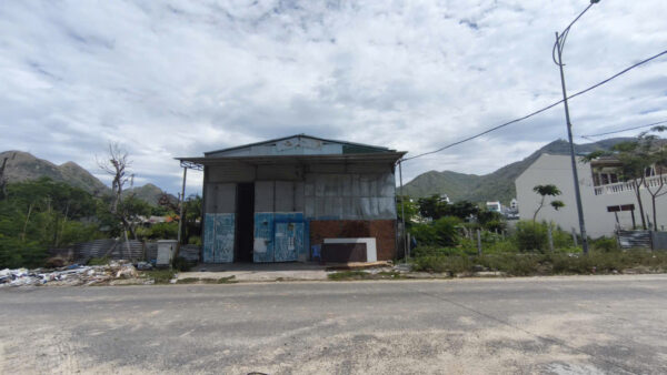 A small, dilapidated warehouse by a road with piles of trash on the left and hills in the distance, under a cloudy sky.