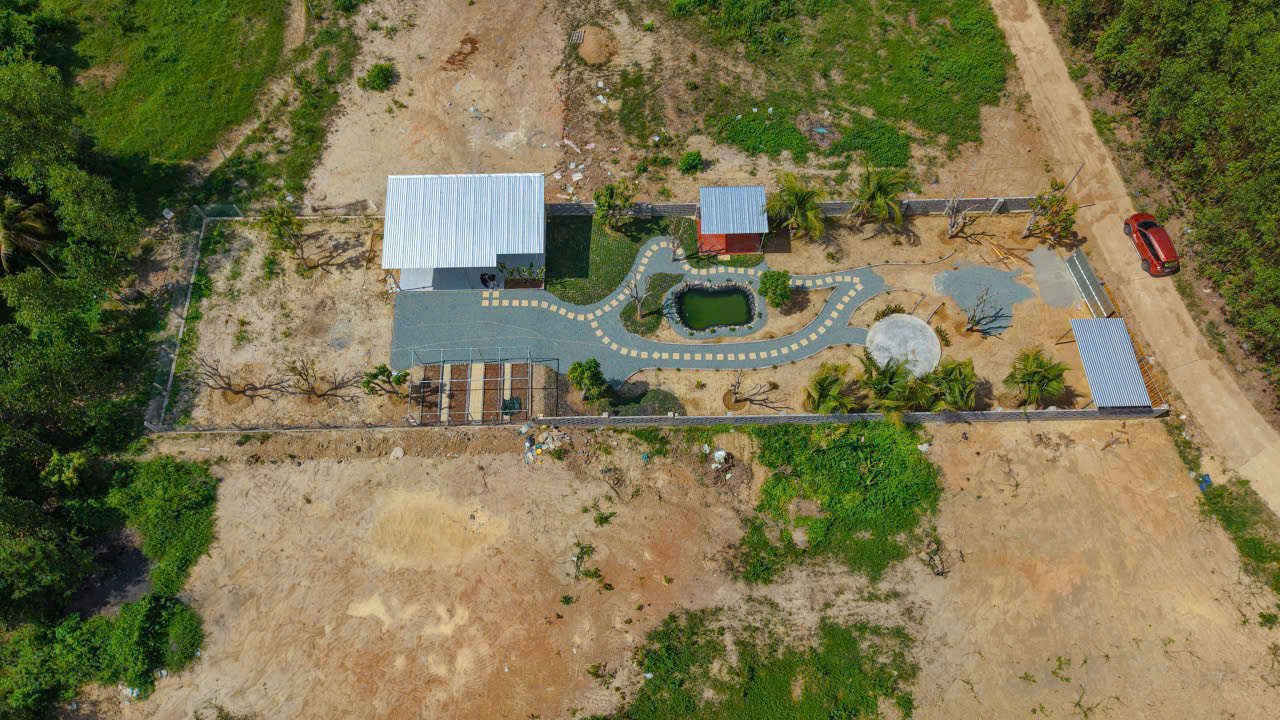 Aerial view of a fenced yard with a winding gray path around a small pond, palm trees, and two metal-roof structures, plus a red shed and a parked red vehicle along a dirt road.