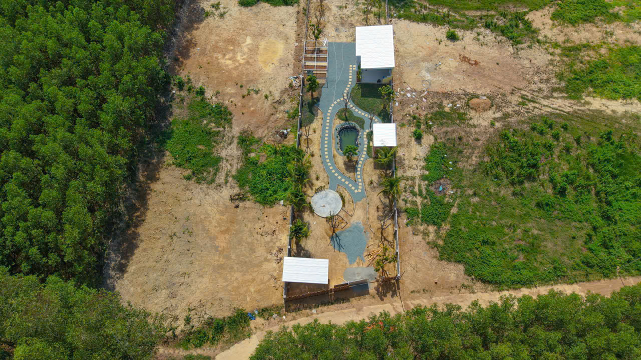 Aerial view of a small fenced park with a winding blue-gray path, a central pond, palm trees, and white-roofed shelters surrounded by dirt and greenery.