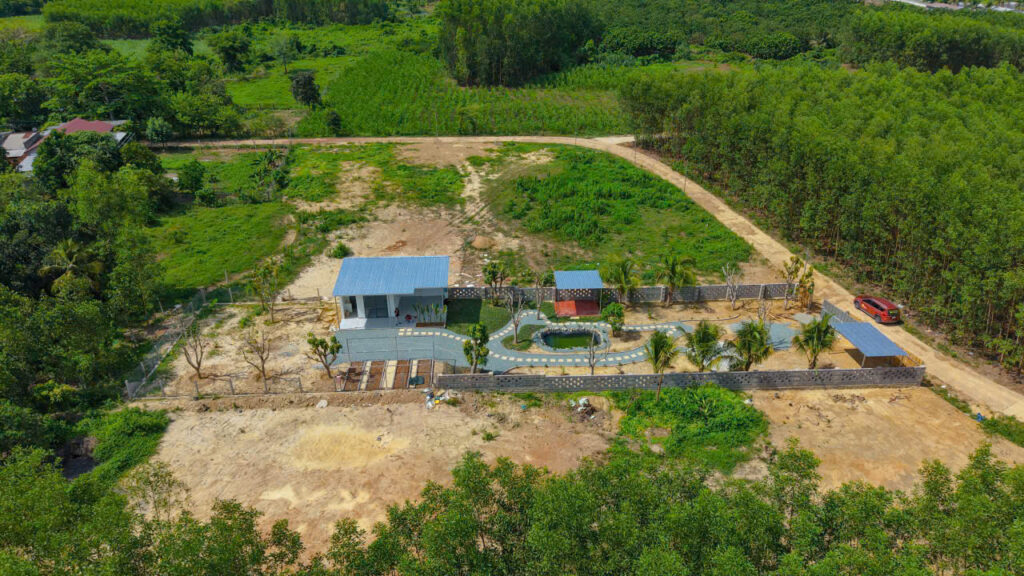 Aerial view of a gated yard with two blue-roof buildings, curved pathway, small pond, palm trees, and a red car along the driveway on the right edge.