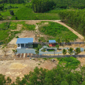Aerial view of a gated yard with two blue-roof buildings, curved pathway, small pond, palm trees, and a red car along the driveway on the right edge.