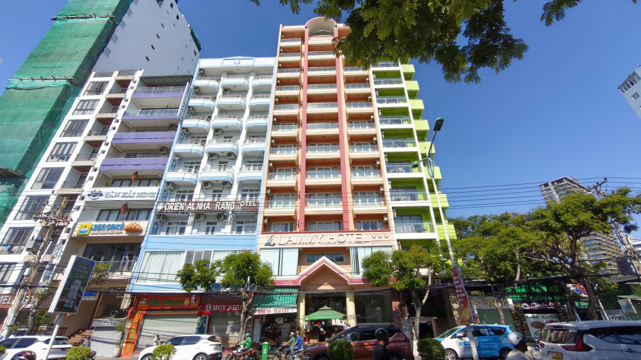 Colorful multi-storey hotel building with pastel facades on a busy urban street, storefronts and parked cars.