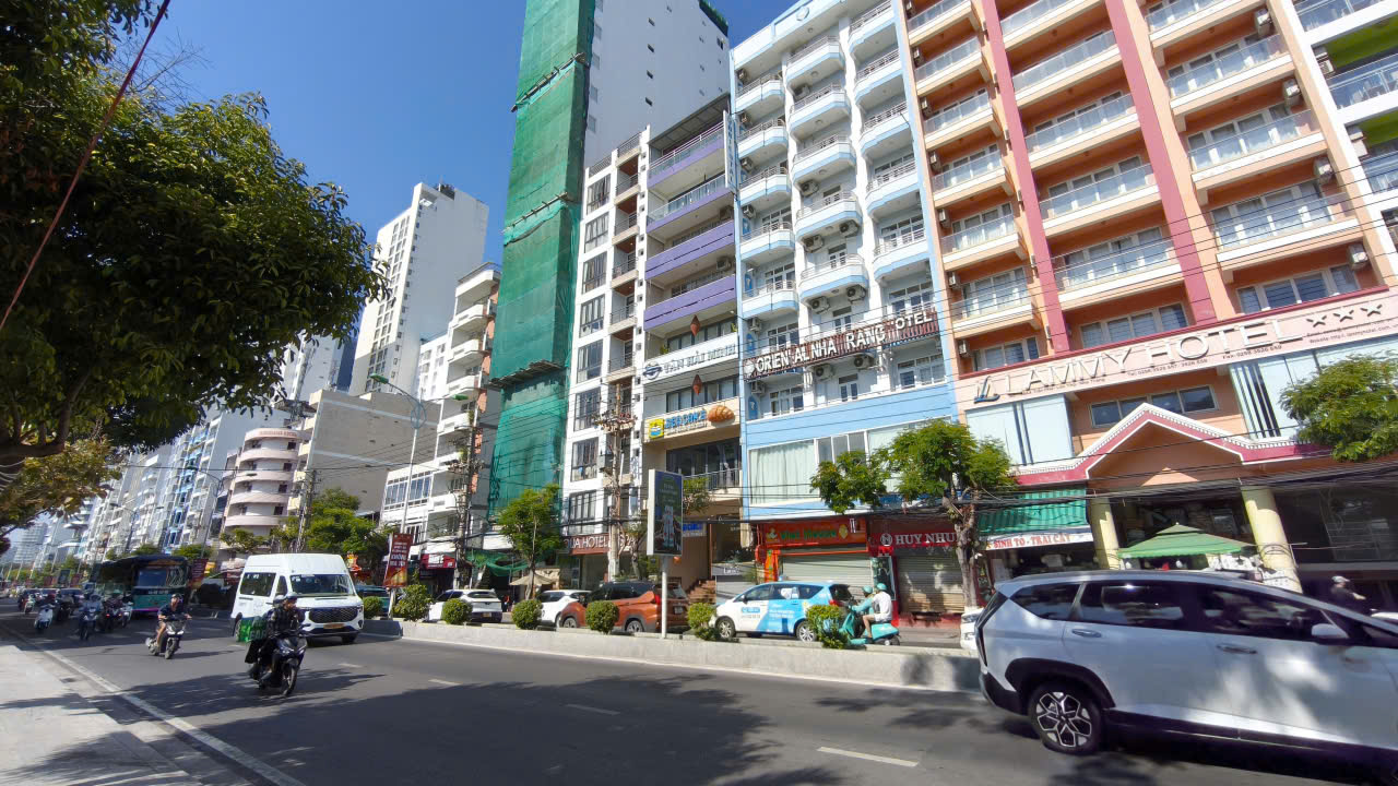 Urban street scene with tall colorful apartment buildings, trees, and parked cars; daytime with light traffic and pedestrians.