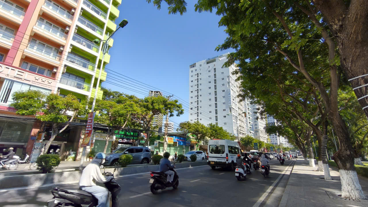 Busy city street with colorful apartment buildings, trees along the sidewalk, and motorcycles and cars traveling in both directions on a sunny day.