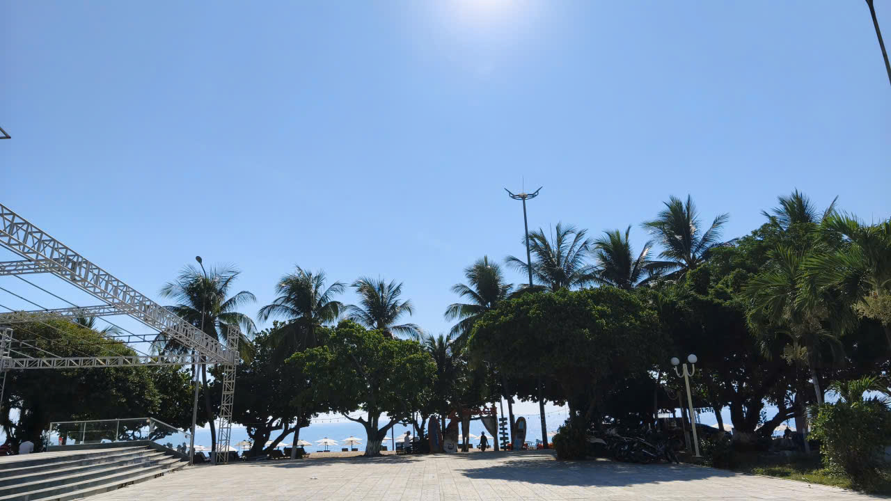Bright sunny day at a beachside promenade with a metal stage frame on the left, palm trees, and the sea visible beyond the trees.